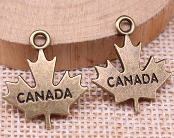 以下が含まれることがあります： 2つのブロンズ色のカエデの葉のチャーム。各チャームには「CANADA」の文字が刻まれ、ジュエリーやその他の工芸品に取り付けるための小さなループが上部にあります。チャームは木製の表面にあります。