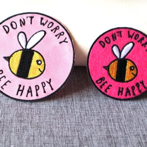 Peut inclure: Deux écussons ronds avec un motif d'abeille. L'un est rose clair, l'autre rose vif. Les deux présentent une abeille jaune et noire et le texte "DON'T WORRY BEE HAPPY" en noir.