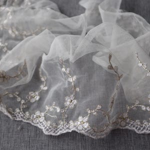 Peut inclure: Tissu de tulle blanc avec broderie florale. Le tissu présente un bord festonné et des motifs floraux détaillés en blanc et marron clair. Idéal pour les projets de mariage ou d'artisanat.