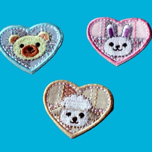 Könnte beinhalten: Drei herzförmige, gestickte Aufnäher mit Tiergesichtern. Ein hellblaues Herz mit einem Teddybären, ein rosa Herz mit einem Hasen und ein beigefarbenes Herz mit einem Welpen. Jeder Aufnäher hat eine dekorative Bordüre.