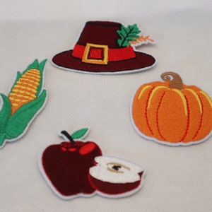 May include: A collection of embroidered patches depicting autumn harvest symbols. Includes a burgundy pilgrim hat, a yellow corn on the cob, an orange pumpkin, and a red apple with a slice. Each patch has a white border.