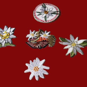 May include: A collection of embroidered edelweiss flower patches. The patches are white, yellow, and green, with detailed stitching. One patch is circular with a red and white border. The background is a solid red.
