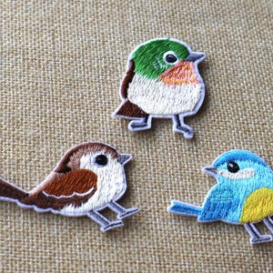 May include: Three embroidered iron-on patches featuring different bird designs. The first bird is green, white, and brown. The second bird is brown and white. The third bird is blue and yellow.