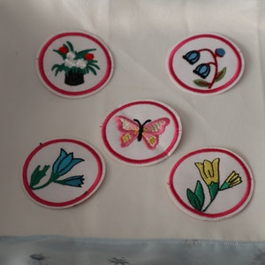 May include: Five embroidered iron-on patches with pink, blue, yellow, green, and white flowers and a butterfly. The patches are circular and have a pink border.