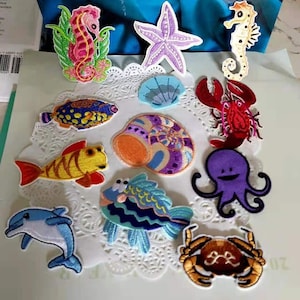 May include: A collection of embroidered patches featuring various sea creatures. Includes a seahorse, starfish, seashell, fish, lobster, octopus, crab, and dolphin. The patches are colorful and detailed, with a variety of textures and patterns.