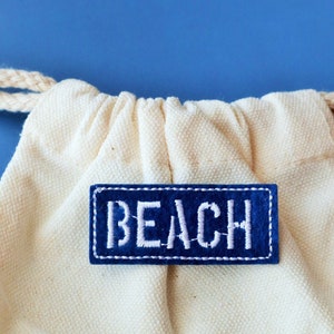 May include: A white canvas drawstring bag with a blue patch that says "BEACH" in white embroidered letters.