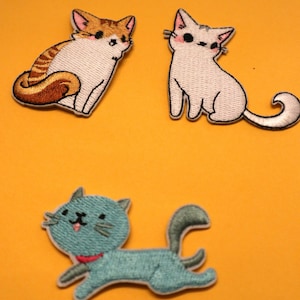 May include: Three embroidered iron-on patches featuring cartoon cats. The first cat is orange and white with a brown tail. The second cat is white with a pink nose and a white tail. The third cat is blue with a red collar and a grey tail.