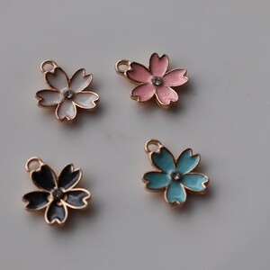 May include: Four gold-tone metal charms shaped like cherry blossoms. Each charm has a different color enamel: white, pink, blue, and black. Each charm has a clear rhinestone in the center.