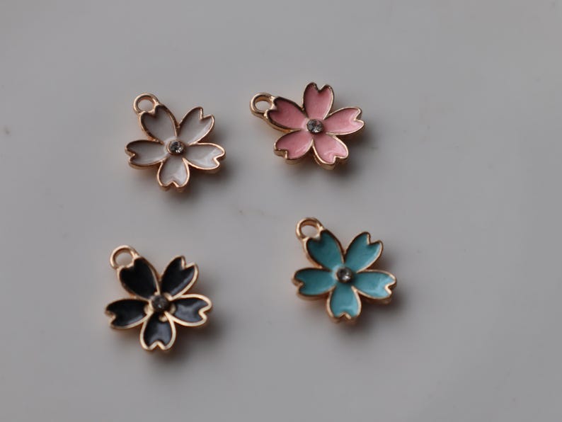 May include: Four gold-tone metal charms shaped like cherry blossoms. Each charm has a different color enamel: white, pink, blue, and black. Each charm has a clear rhinestone in the center.