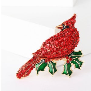 May include: A red cardinal bird brooch with rhinestones and green holly leaves. The cardinal is perched on a branch with red berries.