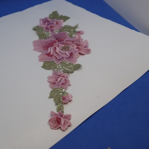 May include: A pink and green floral embroidery applique with a large pink flower in the center and smaller pink flowers cascading down the stem.