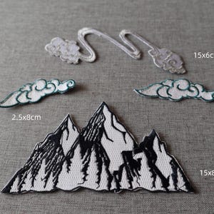 May include: Embroidered patches with mountain and cloud motifs. The mountain patch measures 15cm x 8cm, and the cloud patches are 2.5cm x 8cm and 15cm x 6cm. The patches are white with black and teal detailing, ideal for customising textiles.