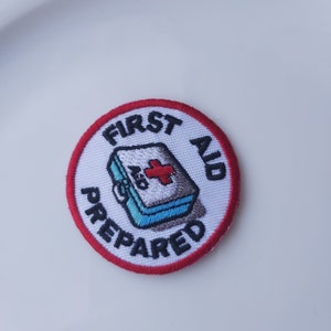 May include: White embroidered patch with a red border. The patch features a first aid kit with a red cross and the text "FIRST AID PREPARED".