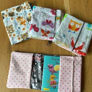 Könnte beinhalten: Vier Stoffbuchhüllen mit verschiedenen Mustern, darunter Eulen, Schmetterlinge und Waldtiere. Eine pinkfarbene Buchhülle mit weißen Punkten und einem Buch mit dem Titel "Drei Monster" ist im Vordergrund.
