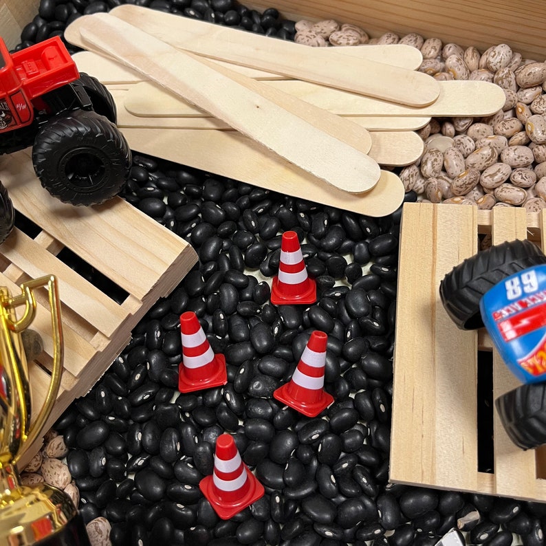 May include: A sensory bin filled with black beans, wooden craft sticks, and toy trucks. Red and white traffic cones are scattered among the beans. A gold trophy is visible in the corner.