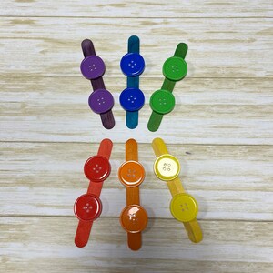 May include: A set of nine wooden craft sticks with colourful buttons attached. The buttons are arranged in a rainbow pattern, with purple, blue, green, yellow, orange, and red buttons.