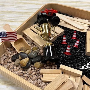 May include: A wooden tray filled with various sensory items. Includes a red monster truck, a gold trophy, small red and white traffic cones, an American flag, wooden blocks, and a mix of beans and rocks. A fun, interactive play set.