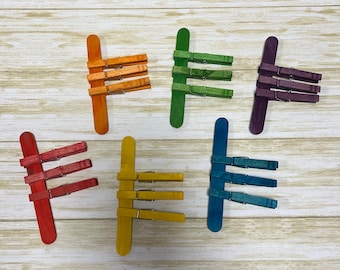 May include: Set of ten colorful wooden clothespins. The clothespins are arranged in a rainbow pattern, with each clothespin a different color: red, orange, yellow, green, blue, indigo, and violet.