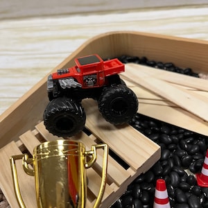 May include: A red and black toy monster truck with large tires, the words "BONE SHAKER" and a skull graphic are visible. A gold trophy and small orange and white traffic cones are also in the scene. The truck is on a wooden ramp.
