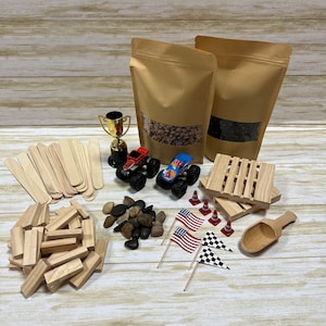May include: An assortment of toys and craft supplies, including two toy monster trucks, wooden blocks, craft sticks, small flags, and a gold trophy. Two brown paper bags with clear windows are also present, along with small rocks and wooden pallets.