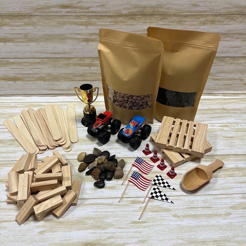 May include: An assortment of toys and craft supplies, including two toy monster trucks, wooden blocks, craft sticks, small flags, and a gold trophy. Two brown paper bags with clear windows are also present, along with small rocks and wooden pallets.