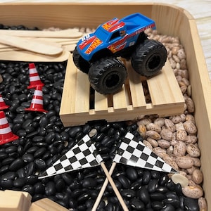 May include: A blue and red Hot Wheels monster truck with the number 68 on the side, parked on a wooden pallet. The scene includes black and brown beans, miniature orange and white traffic cones, and checkered flags.