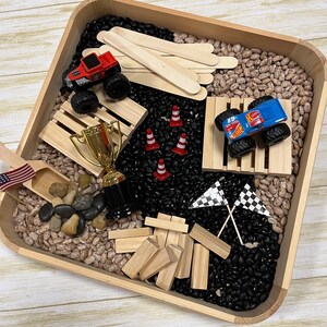 May include: A wooden tray filled with black and pinto beans, toy trucks, wooden blocks, a trophy, miniature traffic cones, and checkered flags. The scene is a sensory play setup, with a small American flag and wooden scoop.