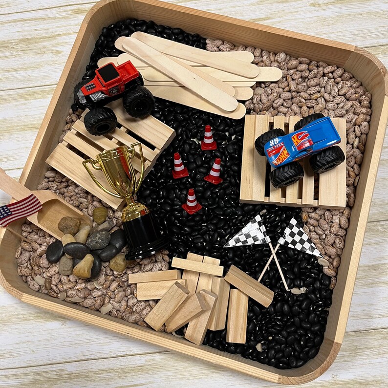 May include: A wooden tray filled with black and pinto beans, toy trucks, wooden blocks, a trophy, miniature traffic cones, and checkered flags. The scene is a sensory play setup, with a small American flag and wooden scoop.