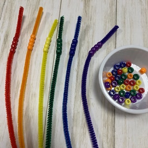 May include: Six colorful pipe cleaners with beads strung on them. The pipe cleaners are red, orange, yellow, green, blue, and purple. A bowl of assorted colorful beads is in the background.