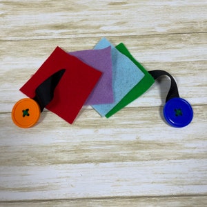 May include: A colorful handmade felt and button craft. The craft features a red, purple, light blue, and green square felt pieces connected by a black ribbon. An orange button is attached to one end, and a blue button to the other.