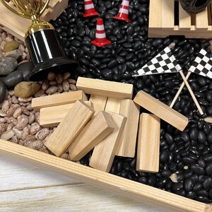 May include: A wooden tray filled with black beans, pinto beans, and small wooden blocks. A gold and black trophy, miniature red and white traffic cones, and checkered flags are also present, suggesting a racing or competition theme.