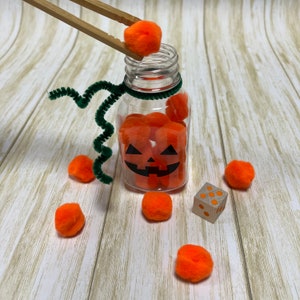 May include: A clear glass jar with a pumpkin face design filled with orange pom poms. A pair of wooden chopsticks are holding another orange pom pom above the jar. There are several orange pom poms scattered around the jar on a white wooden surface.