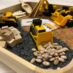 May include: A wooden tray filled with black rice, brown rice, and white beans. There are toy construction vehicles, including a yellow CAT bulldozer, a yellow CAT excavator, and a yellow dump truck. There are also wooden blocks and a small wooden scoop.