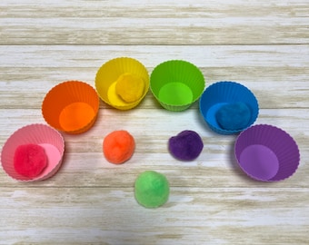 May include: Rainbow-colored silicone cupcake liners with pom-poms in matching colors. The pom-poms are arranged in a semi-circle in front of the cupcake liners.