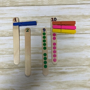 May include: Wooden craft sticks with numbers 1, 2, 9, and 10 printed on them. A blue clothespin is attached to the stick labeled 1. The stick labeled 9 has green dots, and the stick labeled 10 has pink dots and colorful clothespins.