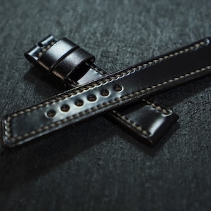 Puede incluir: Dos correas de reloj de cuero negro con costuras blancas, sobre una superficie oscura. Las correas están cruzadas.