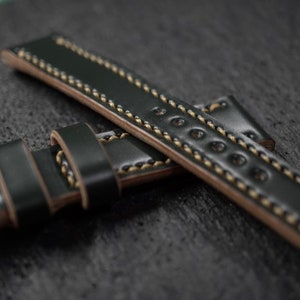 May include: A dark green leather watch strap with a light brown stitching detail. The strap is folded in half, showing the underside of the strap.