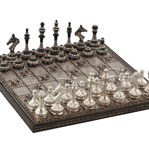 May include: A square chess set featuring a board with alternating dark and light metallic squares. One side has black pieces with thin horizontal metallic bands, while the other side features polished silver-toned pieces with intricate detailing.