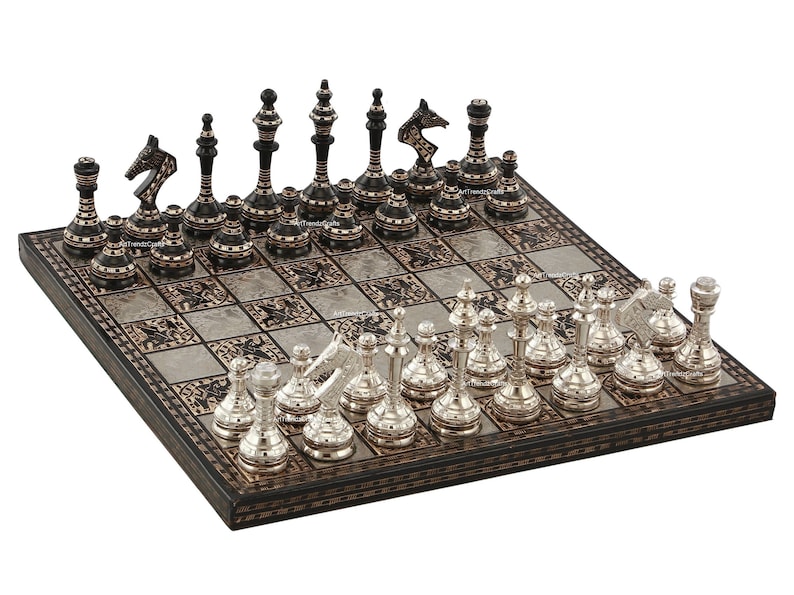 May include: A square chess set featuring a board with alternating dark and light metallic squares. One side has black pieces with thin horizontal metallic bands, while the other side features polished silver-toned pieces with intricate detailing.