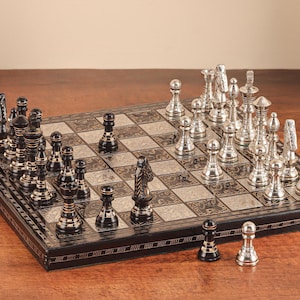 May include: A rectangular chess set featuring a board with alternating black and silver squares, each decorated with intricate patterns. The set includes a collection of polished silver and black metal chess pieces arranged on the board.