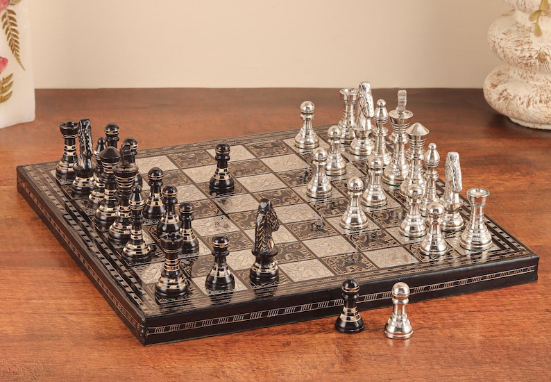 May include: A rectangular chess set featuring a board with alternating black and silver squares, each decorated with intricate patterns. The set includes a collection of polished silver and black metal chess pieces arranged on the board.