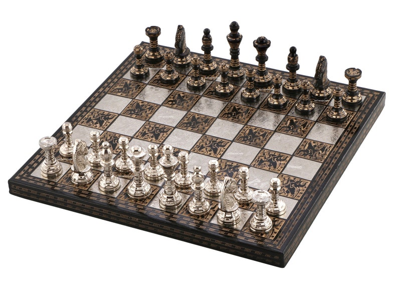 May include: A chess set featuring a square board with alternating silver and black patterned squares. One side has metallic silver chess pieces, while the other side has dark metallic chess pieces with gold-toned accents, all arranged in a standard starting formation.