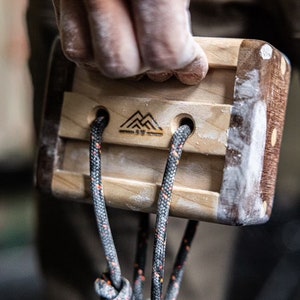 May include: A wooden climbing hold with a textured surface and three holes. The hold is attached to a black rope with orange accents. The hold has a logo of three mountains with the text "317" underneath.