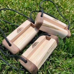 May include: Three wooden blocks with a light wood center and dark wood ends, connected by black cords. Each block has a carved logo. The blocks are resting on green grass.