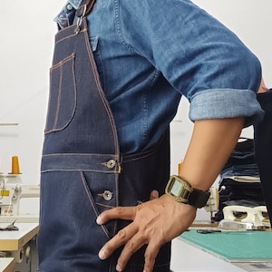 May include: A dark blue denim overall apron with a brown leather strap and a single button closure. The apron has a large pocket on the left side and a smaller pocket on the right side. The person wearing the apron is wearing a light blue denim shirt and a digital watch on their left wrist.