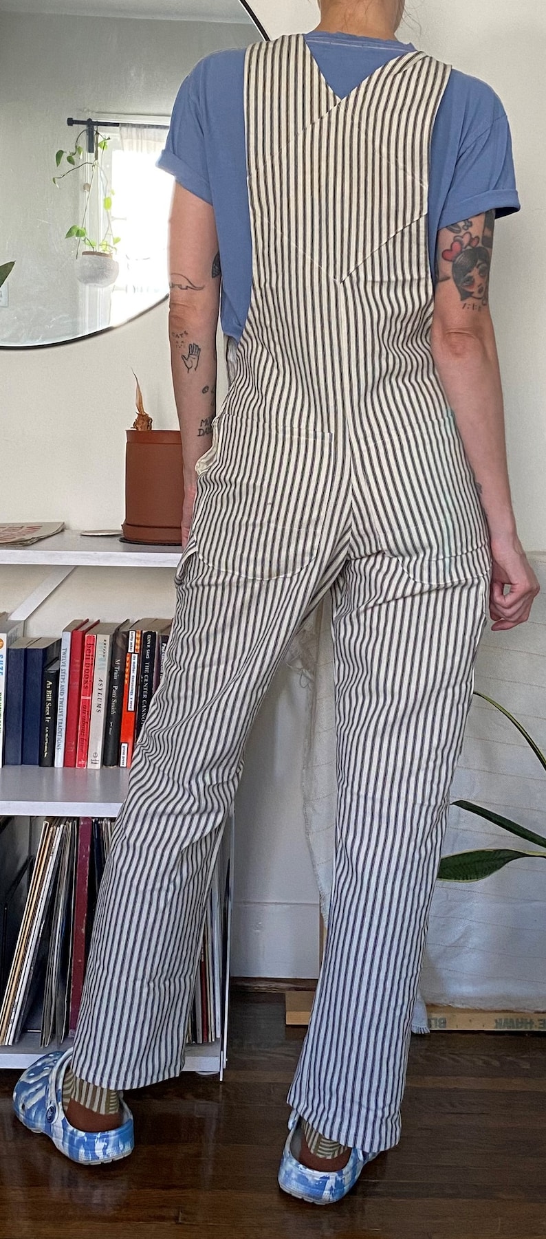 May include: A person wearing a white and black striped overall with a blue t-shirt. The overall has a V-shaped back and two pockets. The person is wearing blue and white patterned clogs.