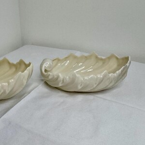 May include: Two off-white, shell-shaped ceramic bowls. Each bowl has a wavy rim and a curled handle on one end. The bowls are placed on a white surface, suggesting a decorative or serving purpose. The design evokes a coastal or nautical theme.