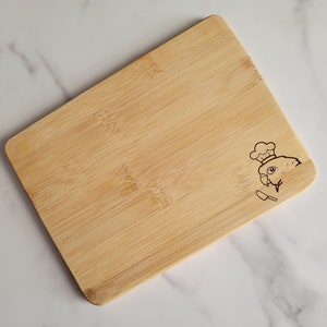 May include: A natural bamboo cutting board with a small, engraved design of a chef's hat and a spatula.