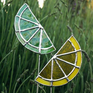 May include: Two stained glass lemon slices, one green and one yellow, on metal stems. The slices are set against a background of green grass.
