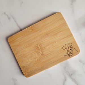 May include: A small, rectangular bamboo cutting board with a laser-engraved design of a chef's hat and a cat holding a knife.